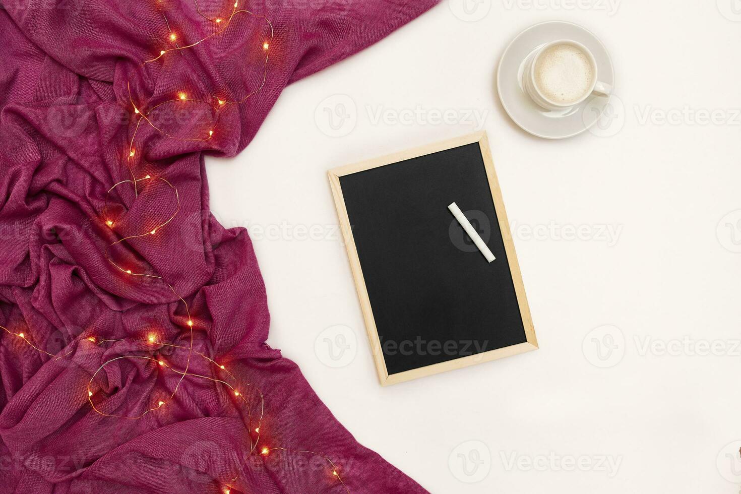 A cup of coffee, a small black wooden board and a white chalk with new years goals. Scarf and Christmas lights on white background. photo