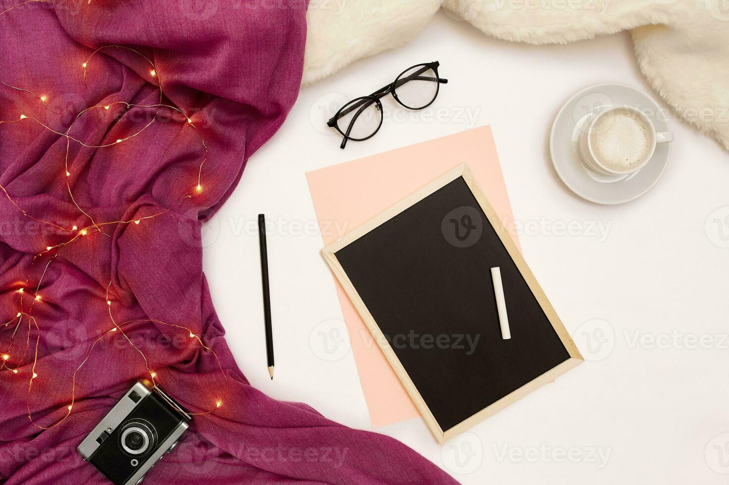 A cup of coffee, a small black wooden board and a white chalk with new years goals. Scarf and Christmas lights on white background. photo