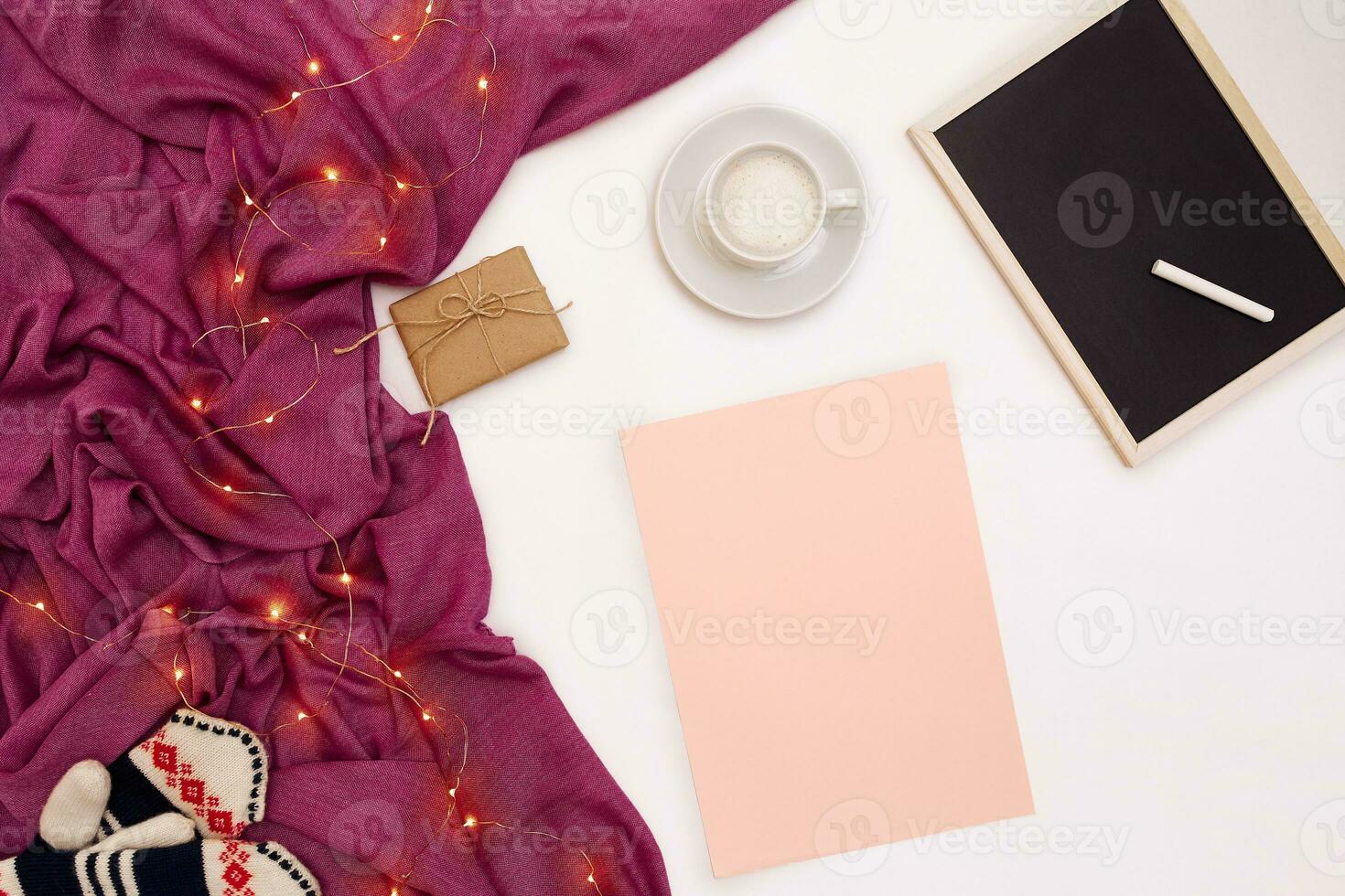 A cup of coffee, a small black wooden board and a white chalk, pink paper with new years goals. Scarf and Christmas lights on white background. photo