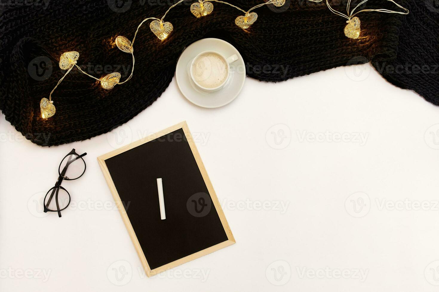 A cup of coffee, a small black wooden board and a white chalk with new years goals. Scarf and Christmas lights on white background. photo