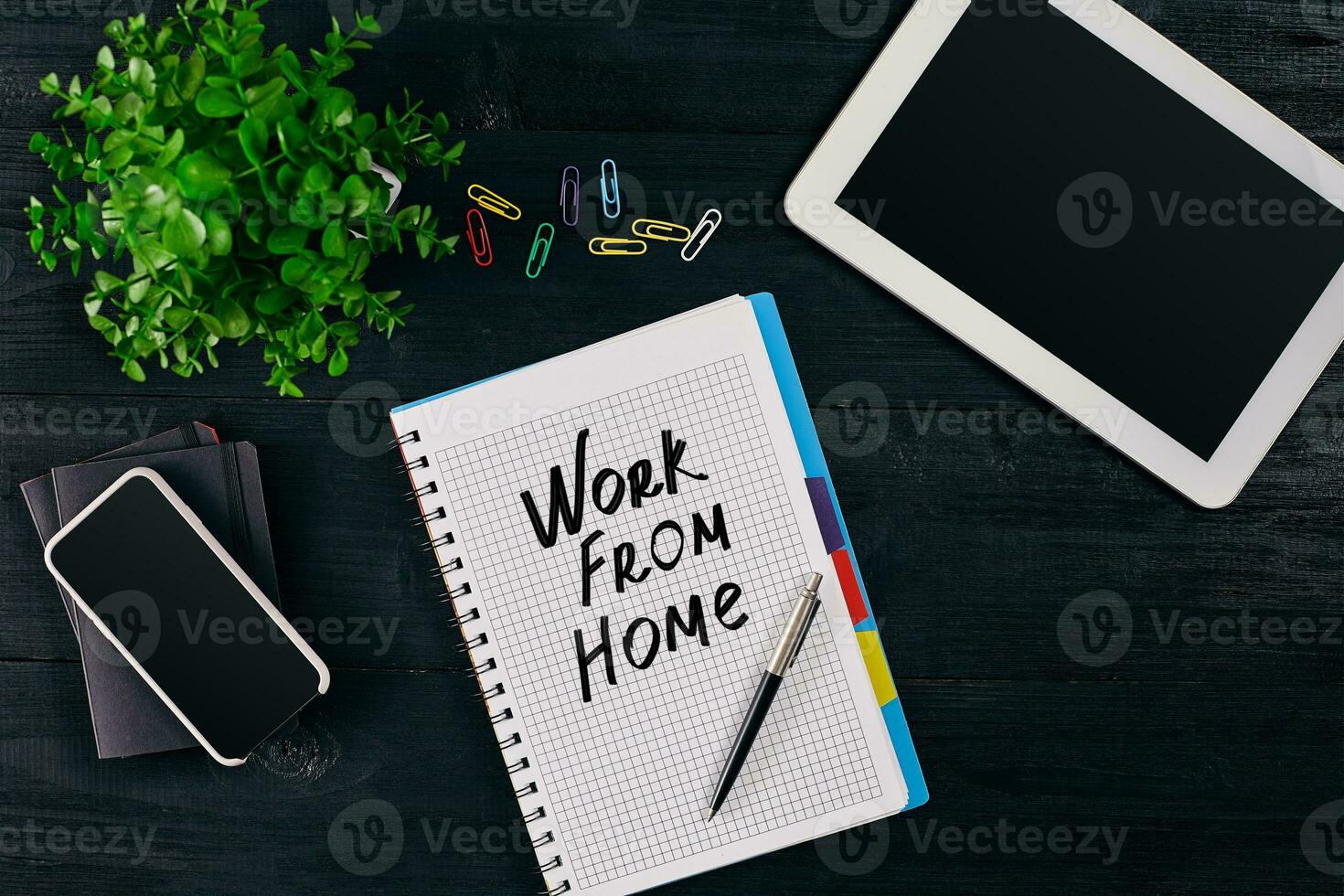 Top view of open notebook written with WORK FROM HOME inscription. Green flower, tablet, colored paper clips, smart, pen and notebook on wooden background. photo