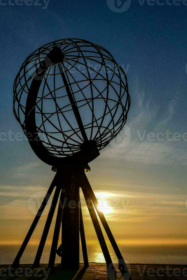 the sun sets behind a large metal sphere in front of the ocean photo