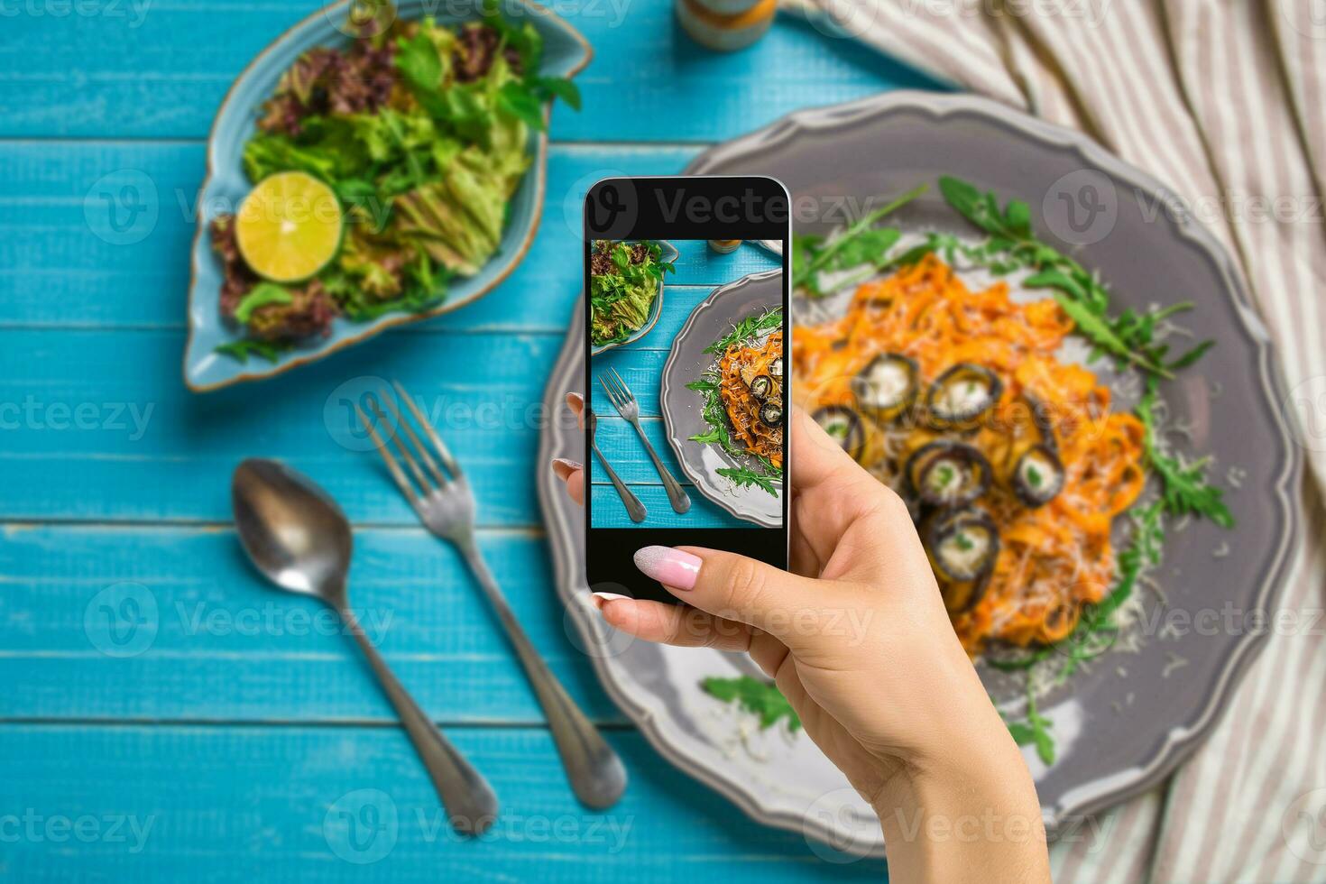 Photographing food concept - woman takes picture of pasta with eggplants, tomato, cheese, arugula and salad photo