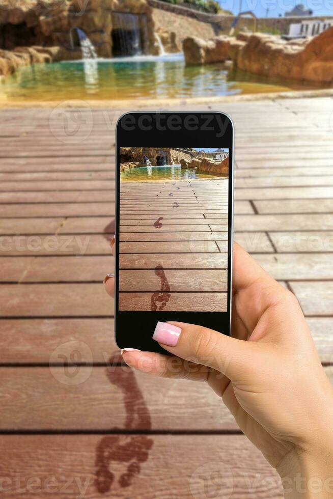 Closely image of female hands holding mobile phone with photo camera mode on the screen. Cropped image of footprints on the wooden floor behind it swimming pool