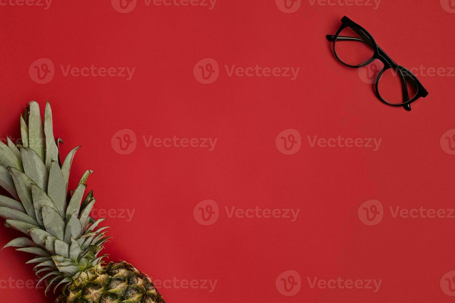 Stylish glasses with black frames and transparent lenses and ripe pineapple against red studio background. Close up, copy space. Top view photo