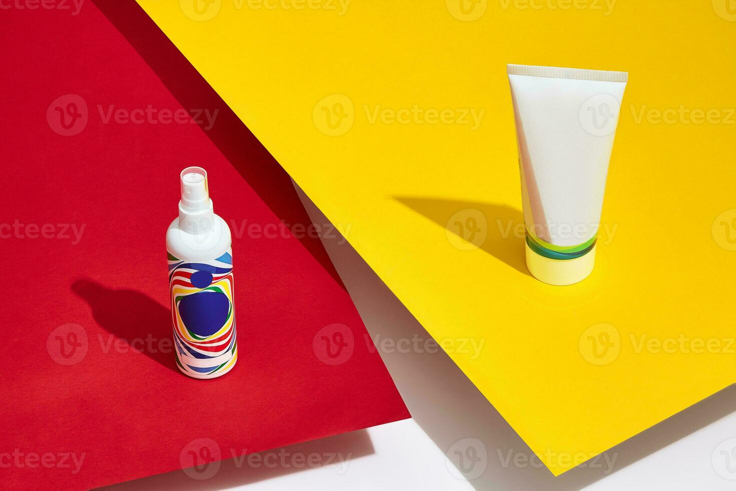 Colorful cosmetic tube and pump bottle with no logo standing on red and yellow sheets of paper above white surface. Close up, copy space photo