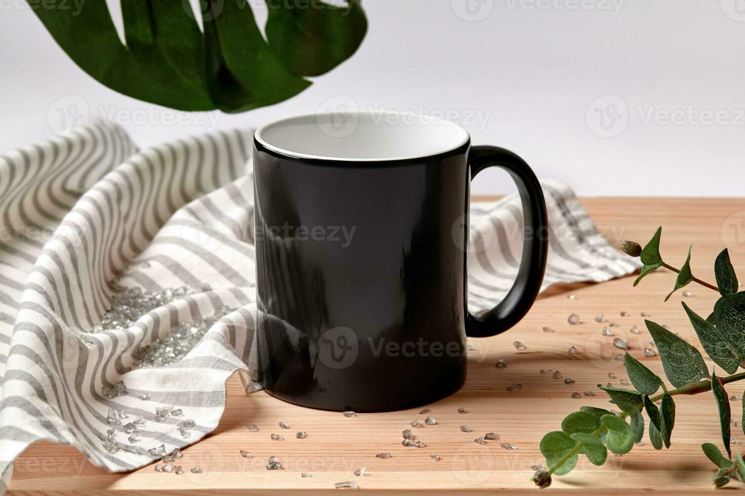 Black porcelain mug on wooden desktop next to striped tablecloth, scattered crystals and green twigs against white background. Close up, copy space photo
