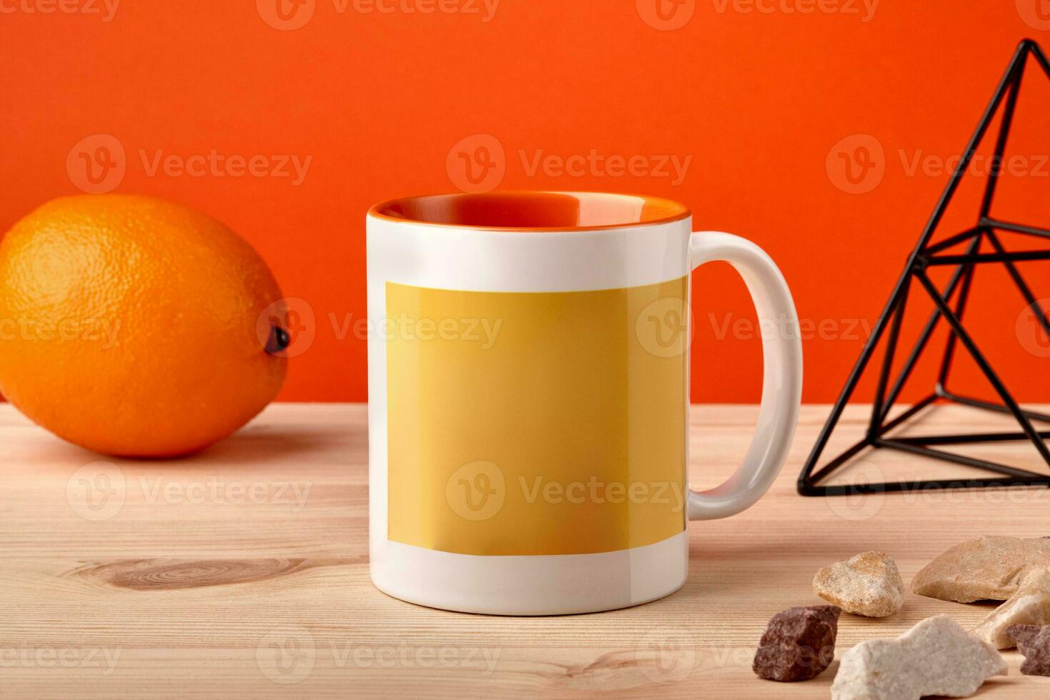 Colorful porcelain mug on wooden desktop next to orange, metal figurine of triangle and small stones against orange background. Close up, copy space photo