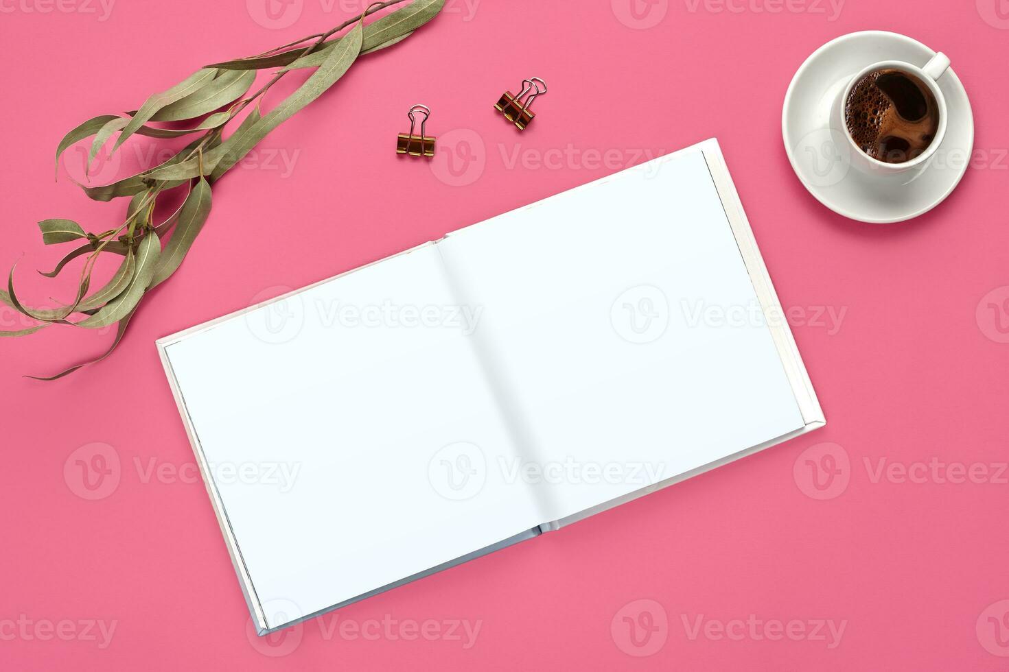 Empty open notebook, two golden binder clips, cup of coffee and green twig on pink background. Stationery, supplies for office. Close up, copy space photo