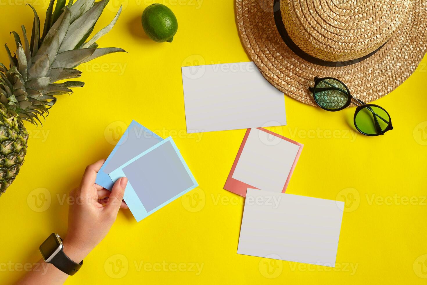 Hand of woman holding snapshot pictures. Straw hat, pineapple, lime, sunglasses and white sheets of paper on yellow background. Close up, copy space. photo