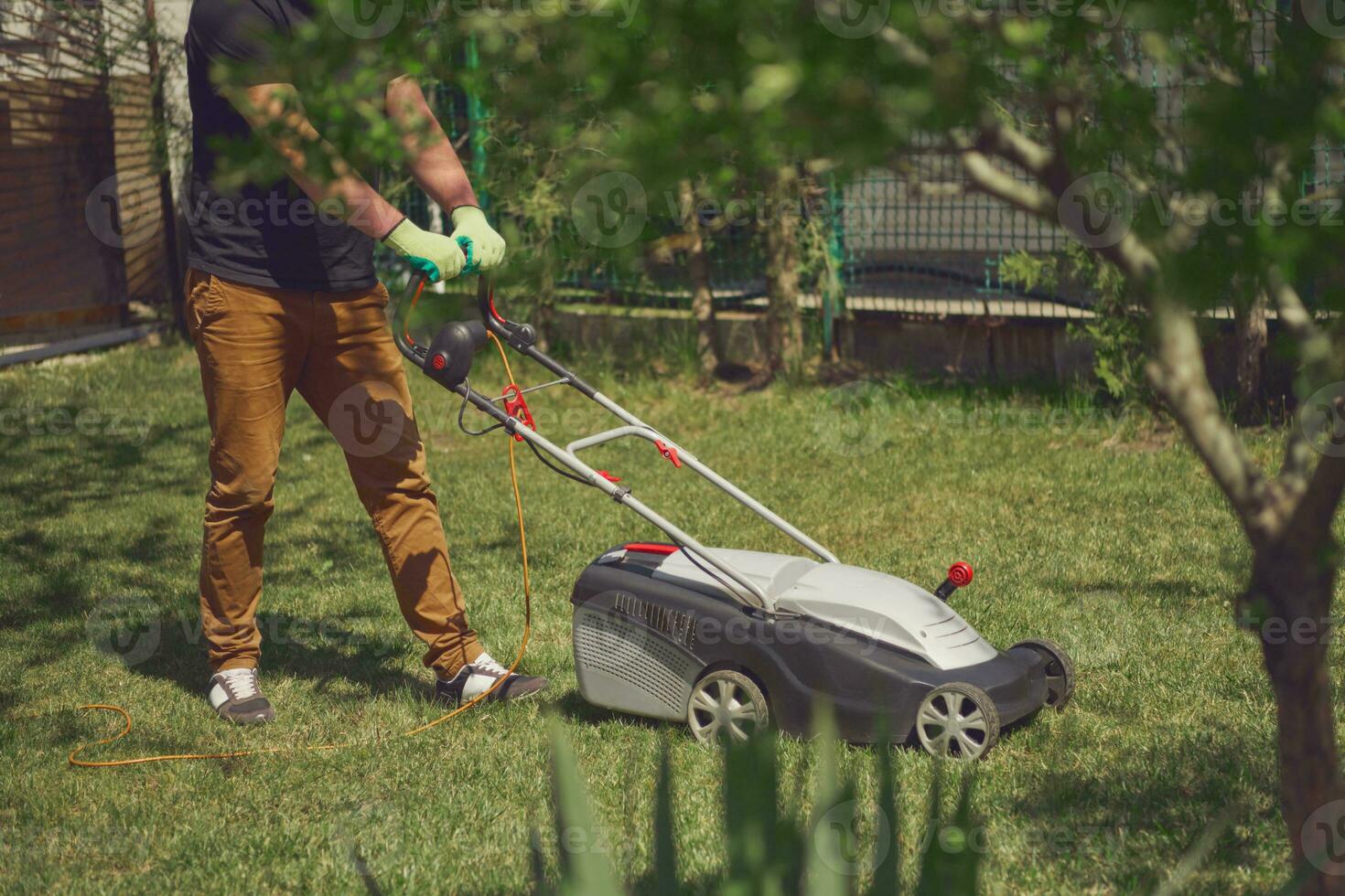 Young worker in casual clothes and gloves is mowing green grass with lawn mower on a yard. Gardening care equipment and services. Sunny day photo