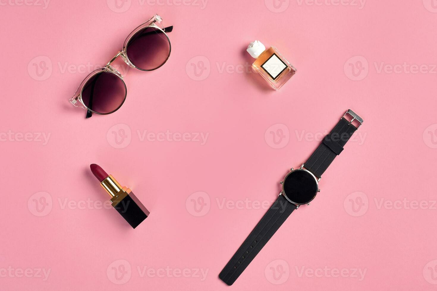 Red lipstick, perfume bottle with blank label, watch with black dial and stylish sunglasses against pink background. Close up, copy space photo
