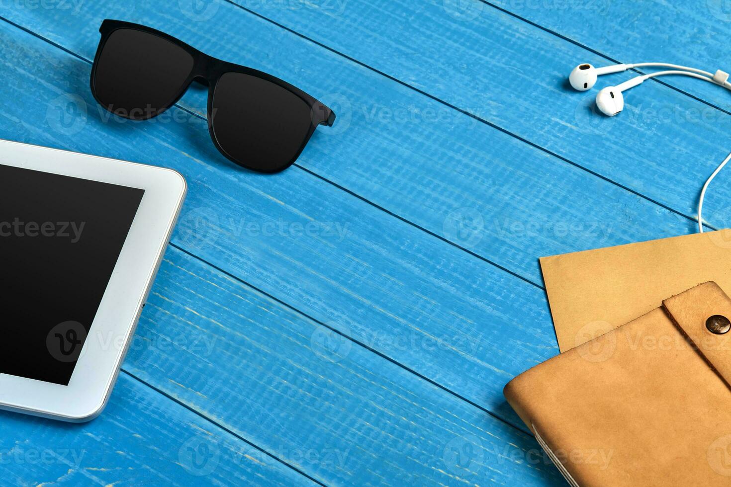 White tablet with black screen, sunglasses, earphones, brown craft paper sheet and leather folder on blue wooden background. Close up, copy space photo