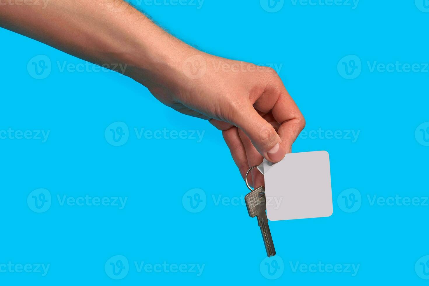 Hand of unknown male is holding a key with blank white square plastic key fob on metal ring against blue studio background. Close up, copy space photo