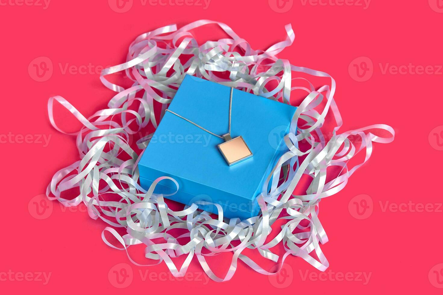 Blank pendant or medallion on a chain on closed blue jewelry box with ribbon against pink studio background. Advertising, gift. Close up, copy space photo