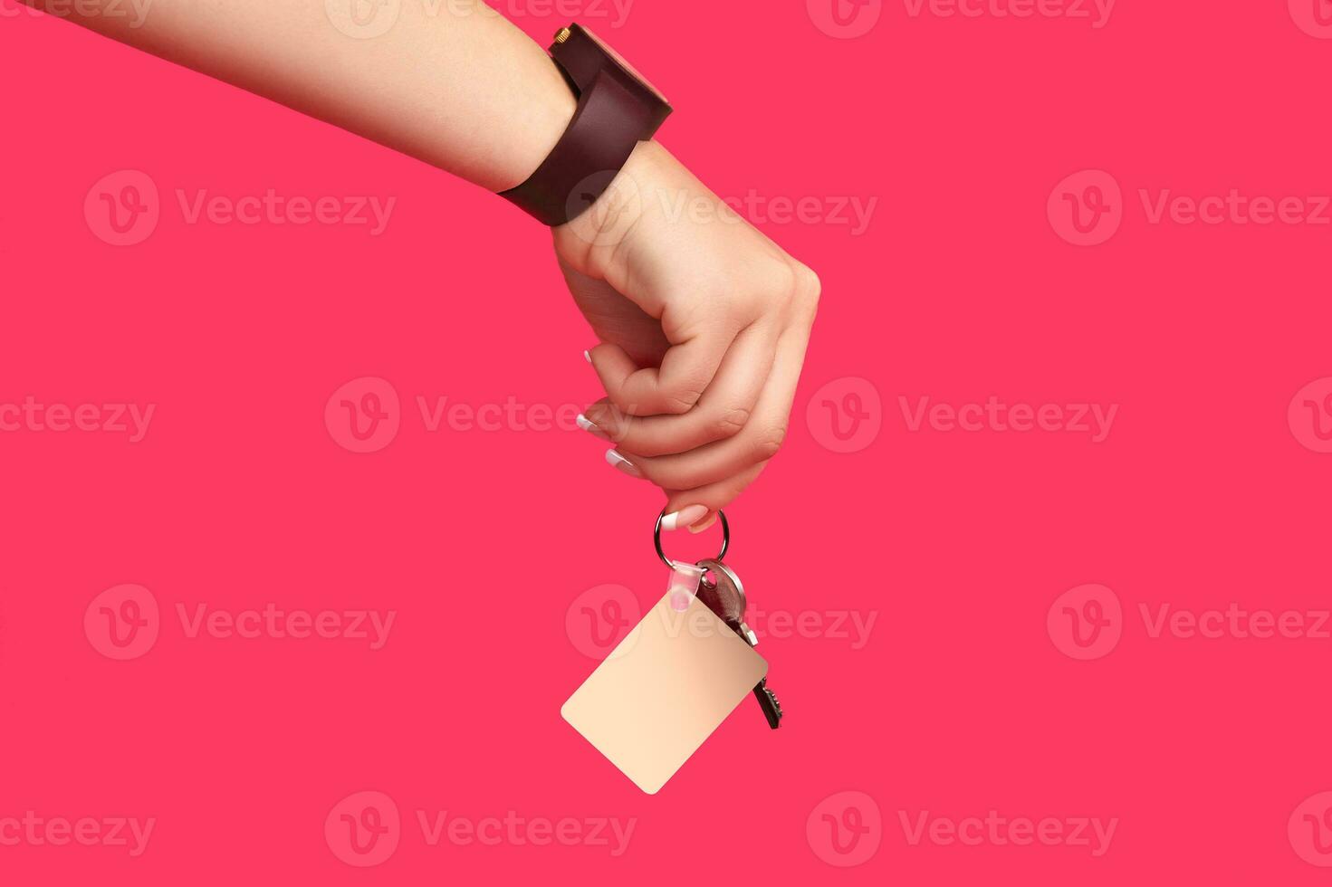 Hand of lady in brown watch is holding a key with blank white square plastic keychain on metal ring against pink background. Close up, copy space photo