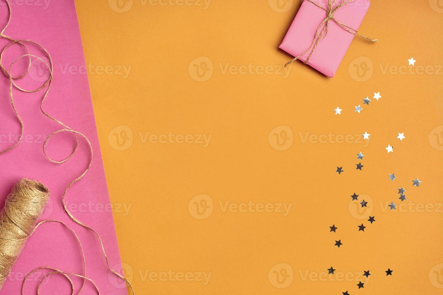 Skein of jute twine or thread, gift box wrapped in pink paper and shiny sequins in form of stars against colorful background. Close up, copy space photo