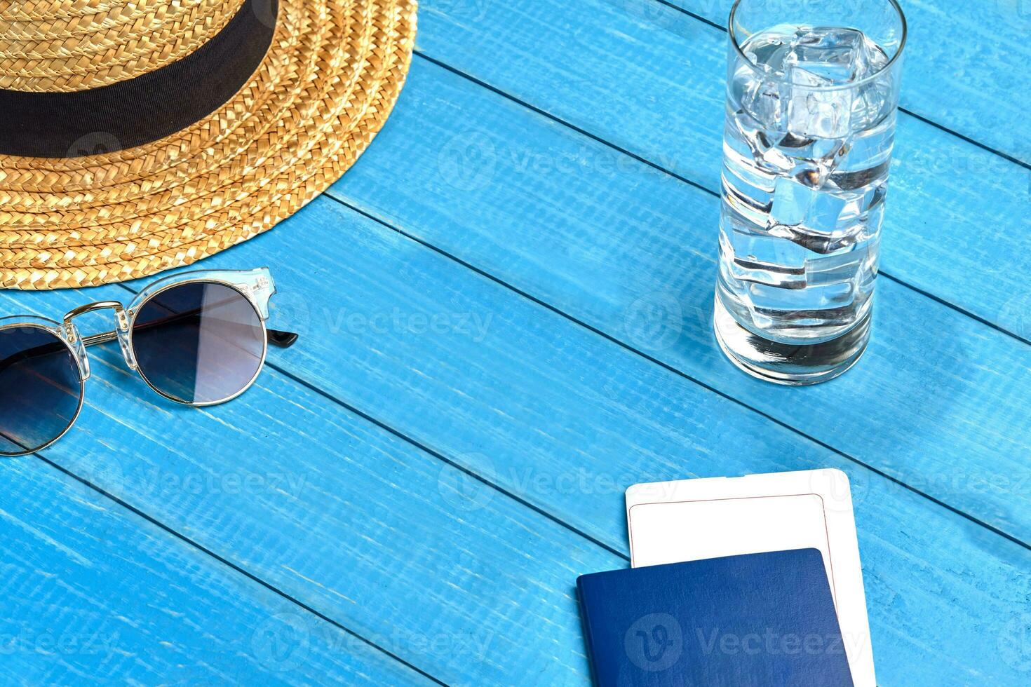 Straw hat, sunglasses, glass of water with ice cubes and passport with ticket on blue wooden background. Travelling and tourism. Close up, copy space photo