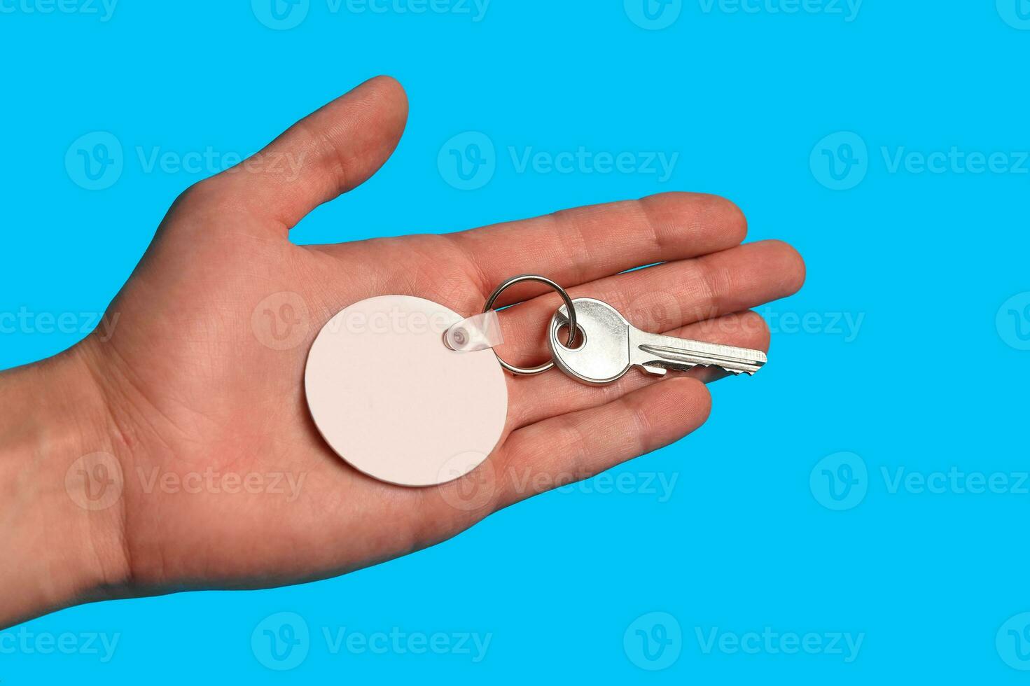 Key with blank white plastic keychain on metal ring on palm of unknown man posing against blue studio background. Close up, copy space photo