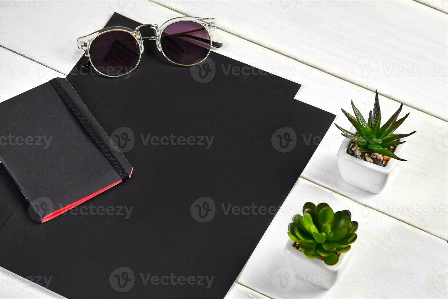 Two black sheets of paper, notebook, sunglasses and green succulents in pots on white wooden tabletop or background. Close up, copy space photo