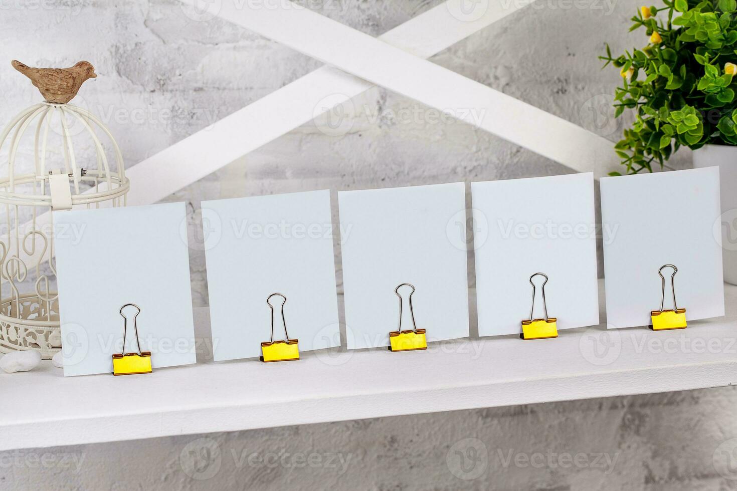 Blank pieces of paper clamped by binder clips on white shelf next to artificial green plant in pot and bird sitting on cage. Close up, copy space photo