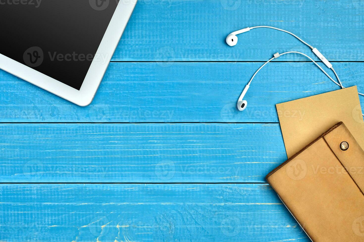 White tablet with black screen, earphones, brown craft paper sheet and leather folder on blue wooden desktop. Close up, copy space photo