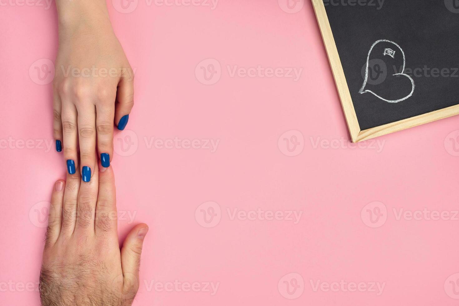 Hand of unknown female is touching a mans one against pink background, next to a small chalkboard with heart drawn on it. Close up, copy space photo