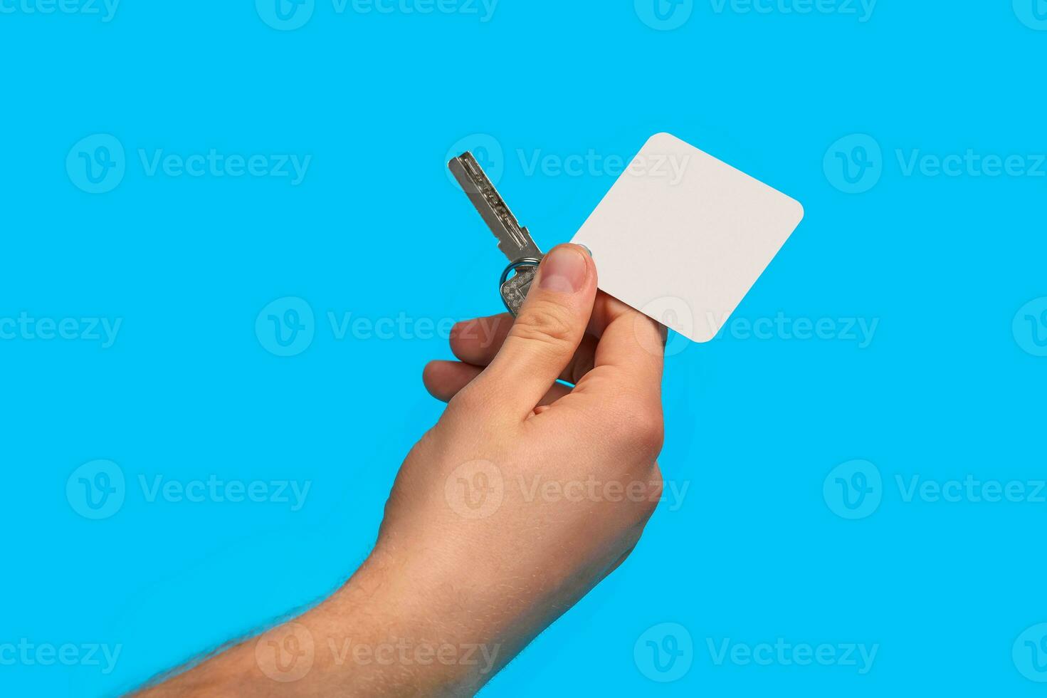 Hand of unrecognizable guy is holding a key with empty white square plastic key fob on metal ring against blue studio background. Close up, copy space photo