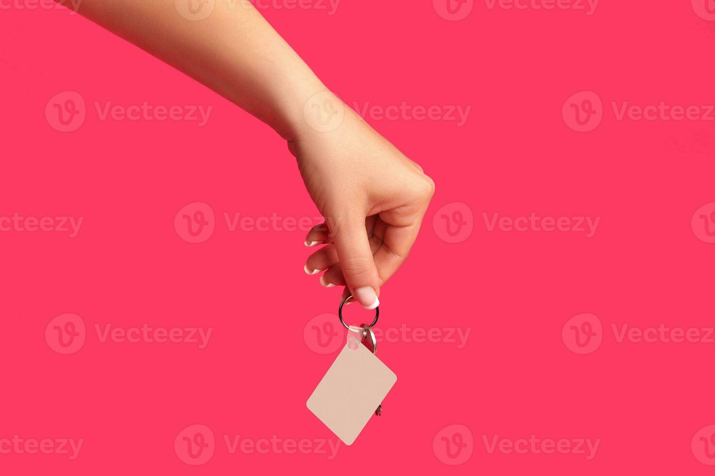 Hand of unrecognizable female is holding a key with empty white square plastic key fob on metal ring against pink background. Close up, copy space photo