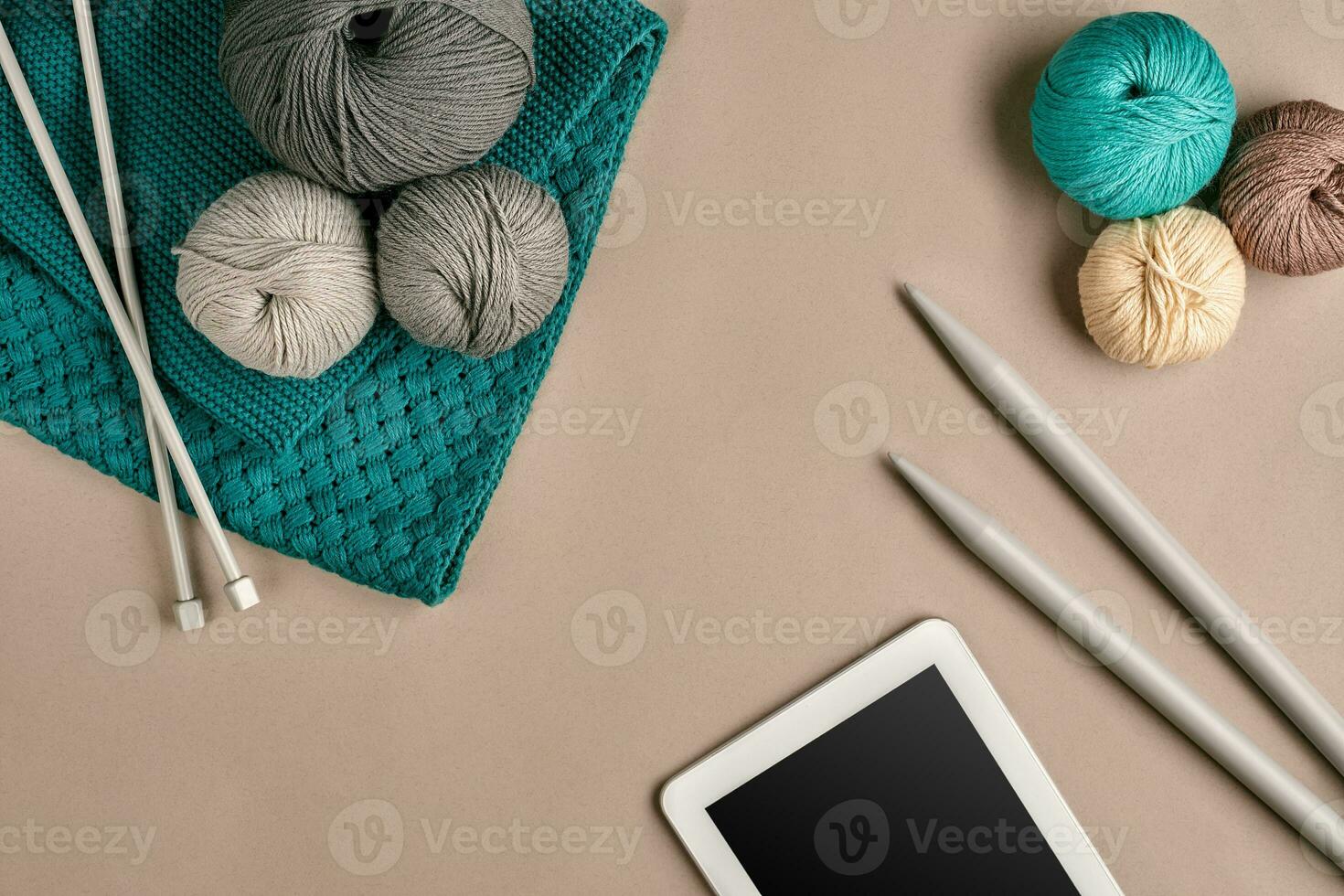 Grey and turquoise knitting wool, knitting needles and a tablet with a black screen on beige background. Top view. Copy space photo