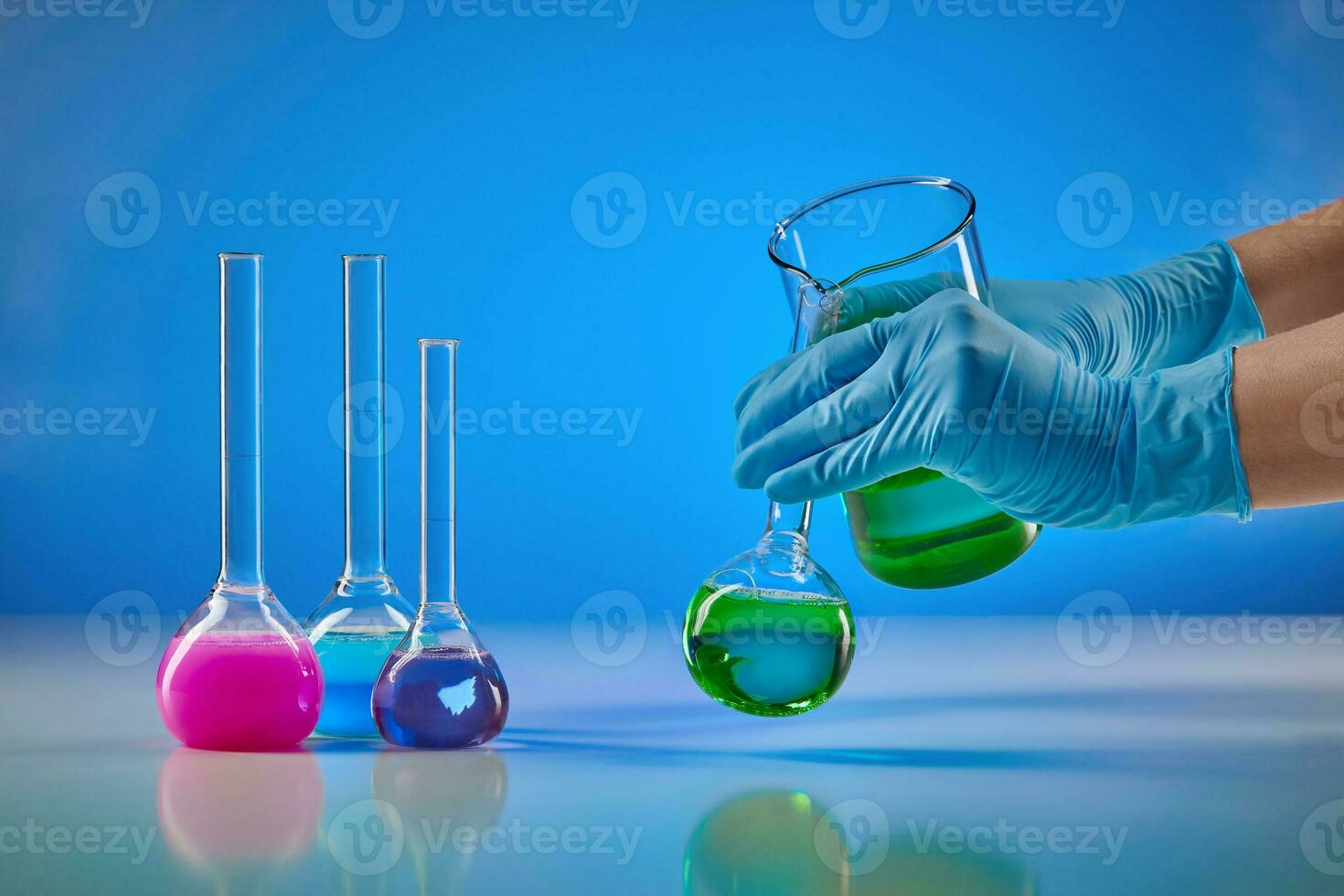 Hands in disposable gloves pouring green liquid from beaker into medical flask. Colorful chemical reagents, blue background. Pandemic of Coronavirus photo