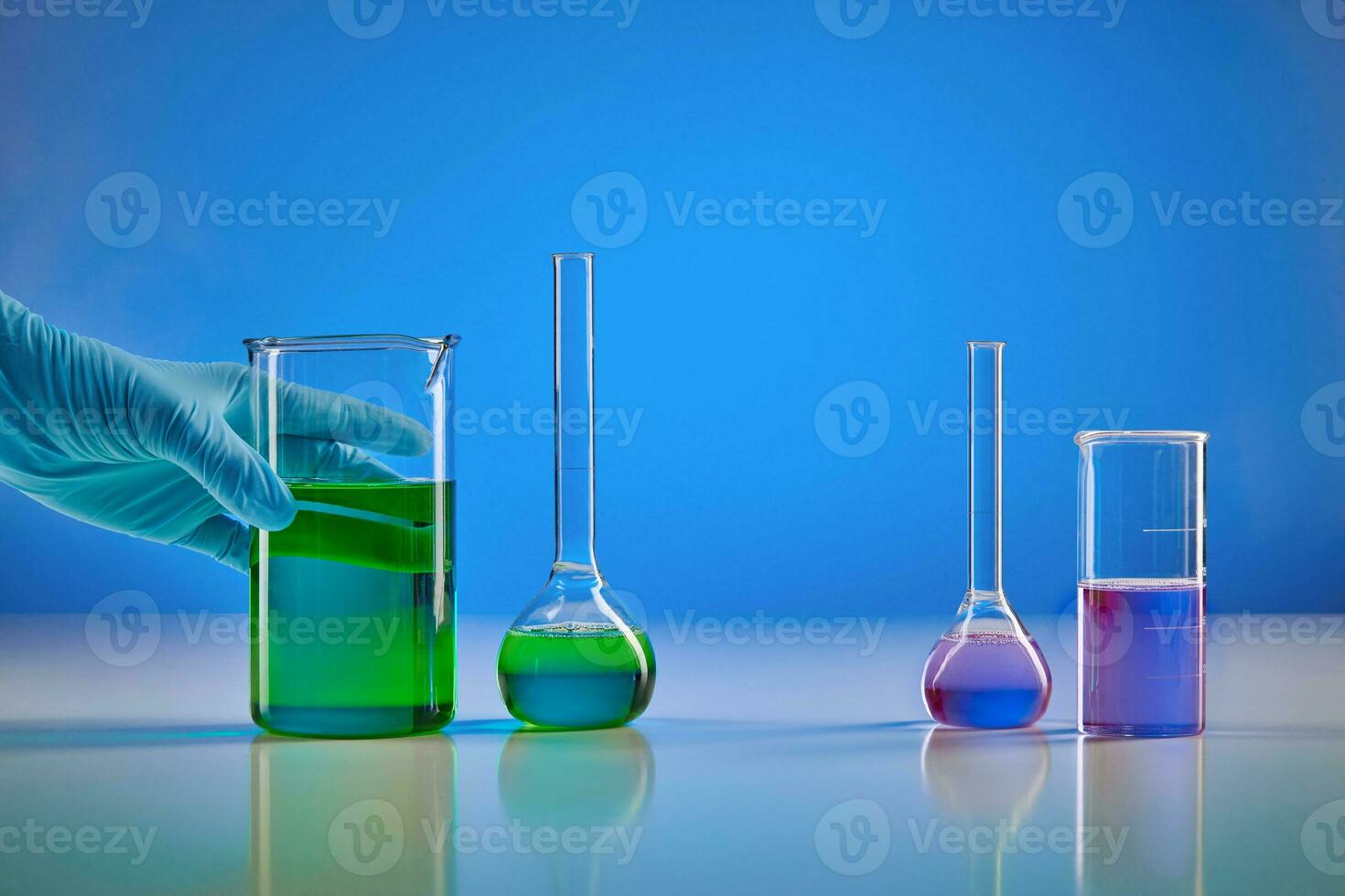 Hand in disposable glove holding beaker, green liquid. Medical flasks, purple chemical reagents, blue background. Coronavirus, pandemic COVID-19 photo