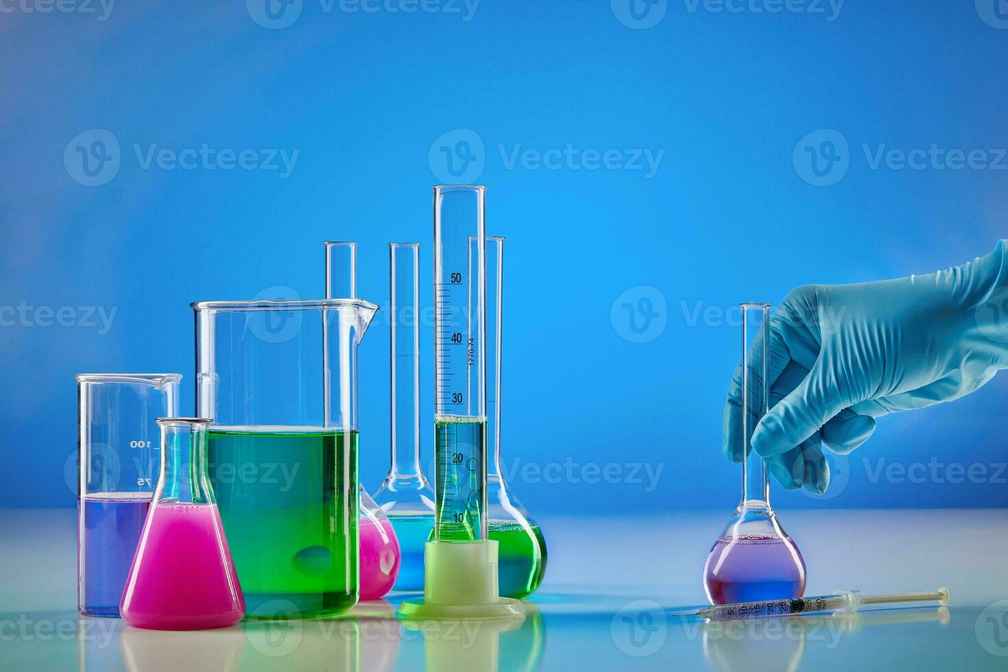 Hand in disposable glove holding medical flask. Colorful chemical reagents in beakers and measuring cylinder, blue background. Coronavirus pandemic photo