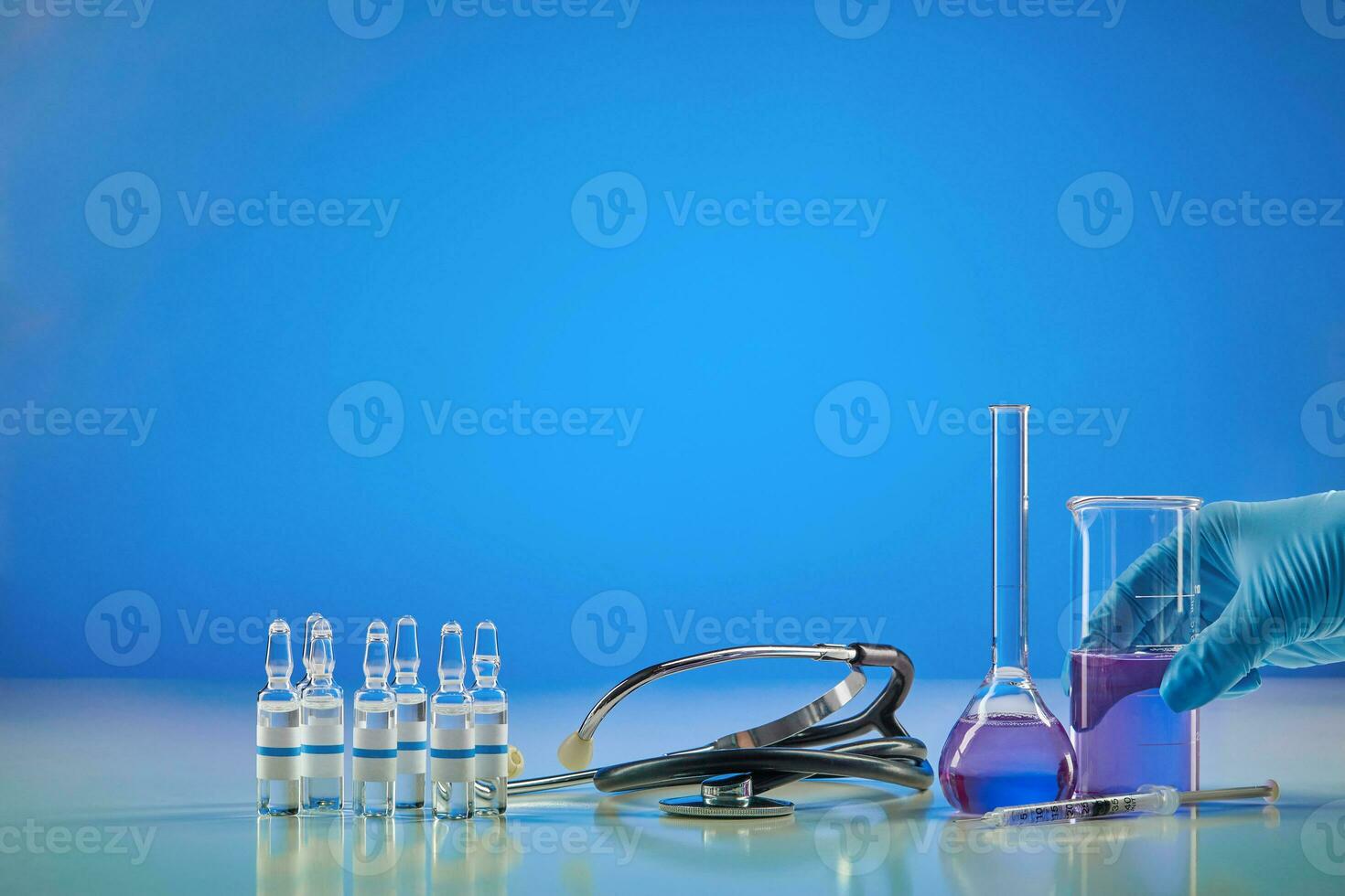 Coronavirus. Hand in glove holding beaker with purple reagent. Six ampoules with liquid, syringe, medical flask and phonendoscope, blue background. photo