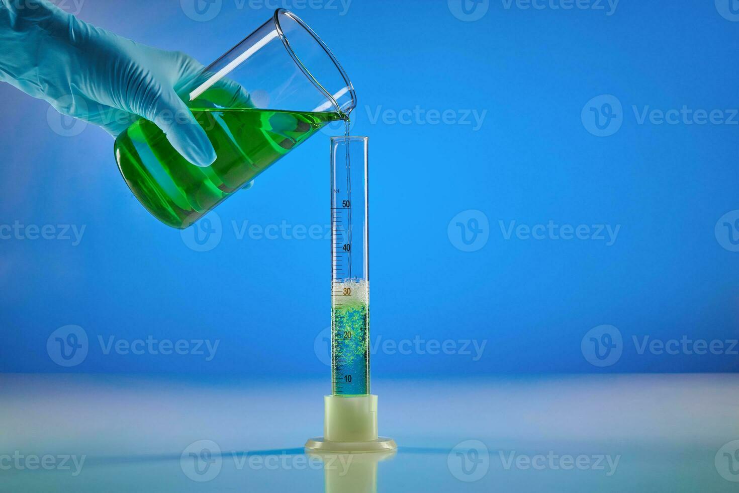 Hand in disposable glove is pouring green liquid from beaker into measuring cylinder, blue background. Coronavirus. Worldwide pandemic COVID-19 photo