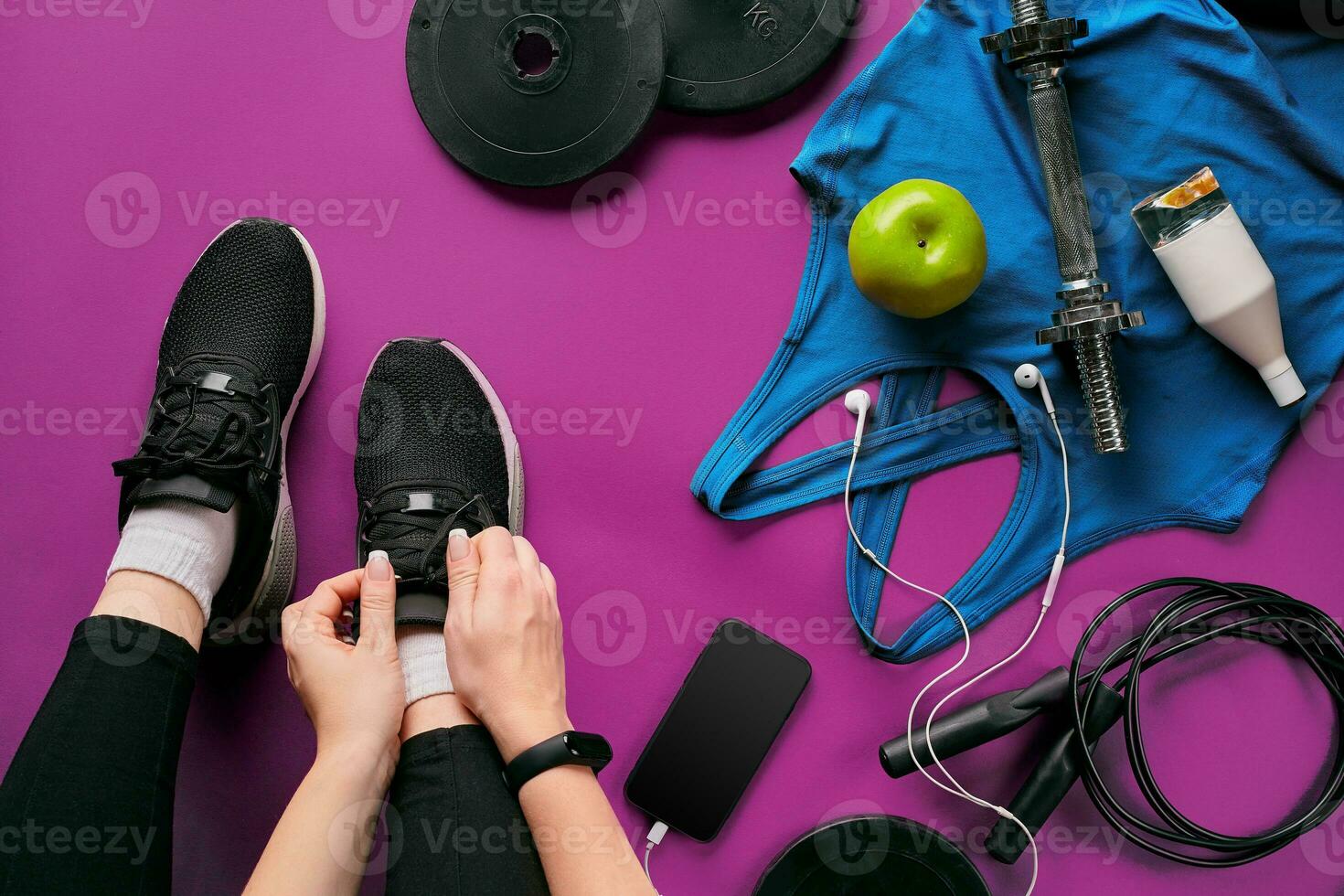 Young woman laces sneakers, preparing for training. Bottle of water, yoga mat, phone, headphones on purple background flat lay top view. photo