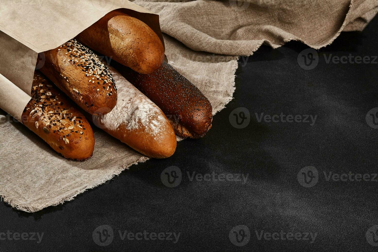 Variety of fresh baked bread in a paper bag, laying on a burlap against black background, copy space. Close-up photo