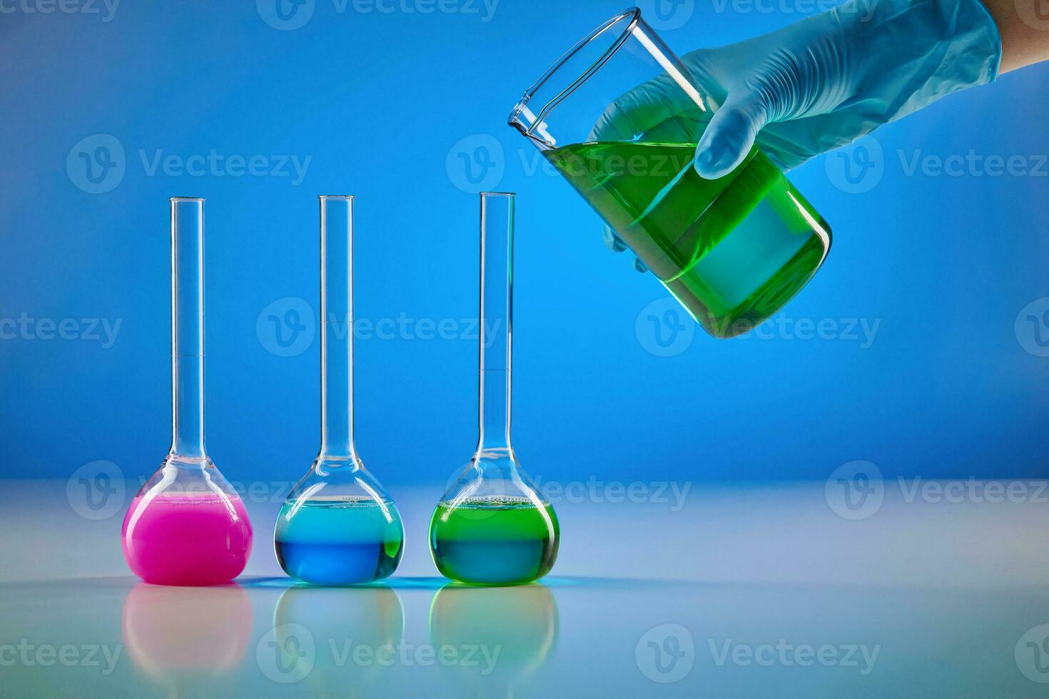 Hand in disposable glove is pouring green liquid from beaker into medical flask. Colorful chemical reagents, blue background. Pandemic of Coronavirus photo