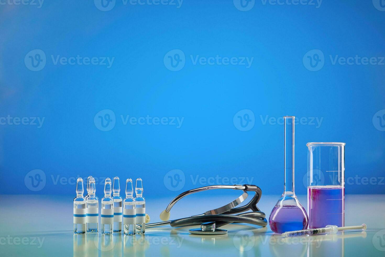 Beaker, medical flask with purple chemical reagent. Six ampoules with liquid, syringe and phonendoscope, blue background. Worldwide pandemic COVID-19 photo