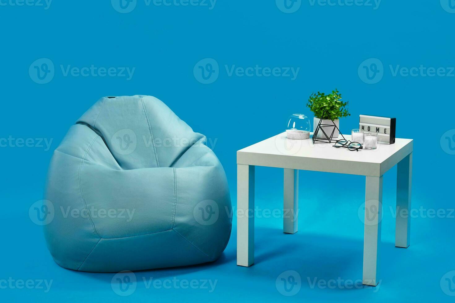 Bean bag, white coffee table with candles on it and in glass candlestick, green flower in pot, glasses, decorative iron triangle. Blue background photo