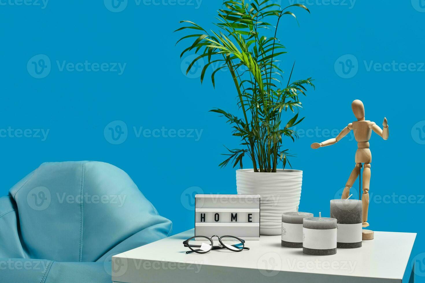 Turquoise armchair bean bag, white coffee table with green flower in pot, three candles, wooden figurine of man, glasses. Blue background. Close up photo