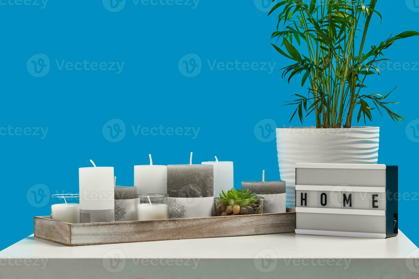 White table with green plant and succulent in pots, different sized candles in wooden stand, lamp with inscription home. Blue background. Close up photo