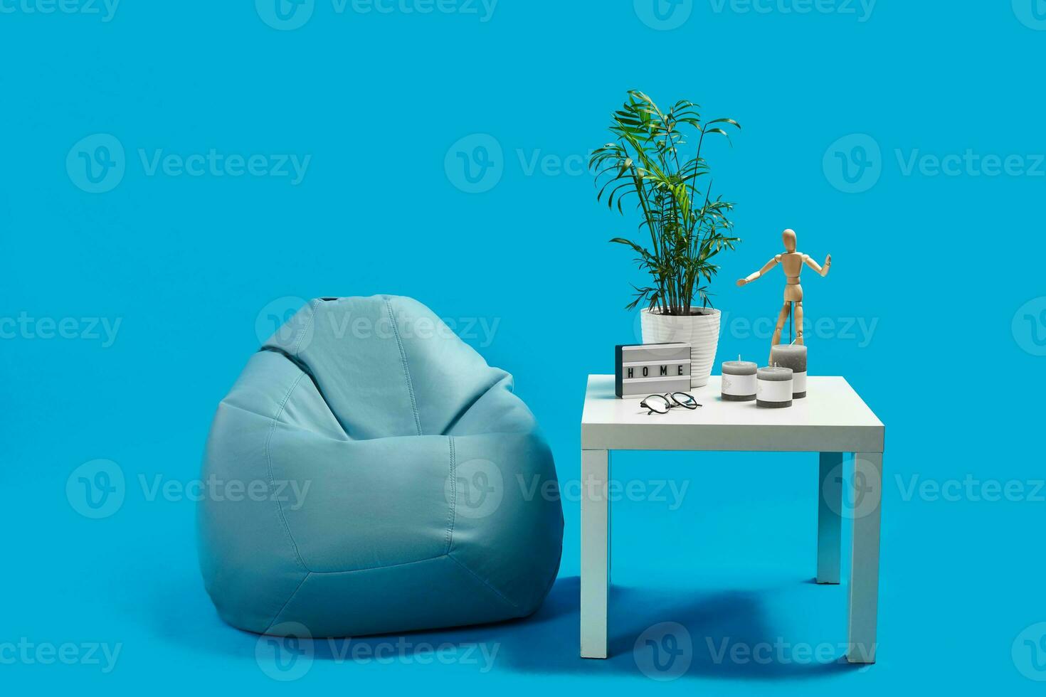 Turquoise bean bag, white coffee table with green flower in pot, three candles, wooden figurine of human, glasses. Blue background. Copy space photo