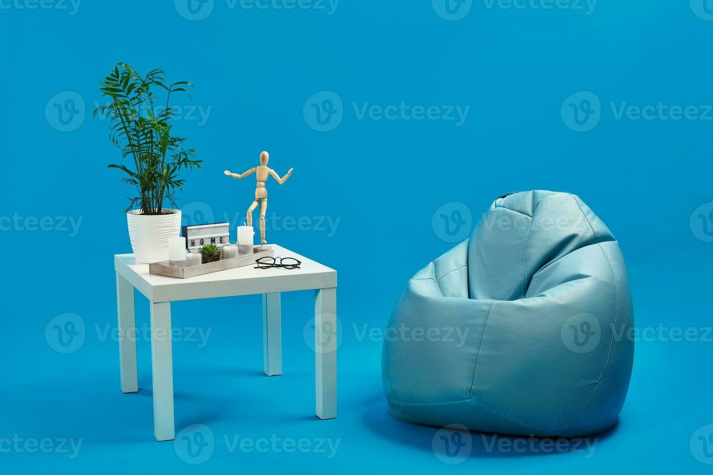 Turquoise bean bag, white coffee table with green flower in pot, candles, succulent, wooden figurine of human, glasses. Blue background. Copy space photo