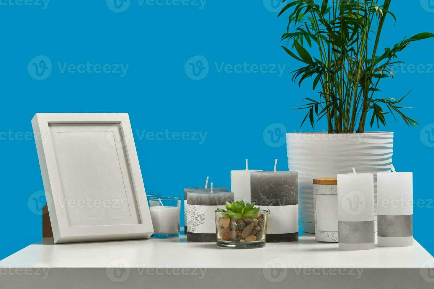 White table with green plant and succulent in pots, some candles, empty photo frame, jar with a wooden lid and copy space. Blue background. Close up