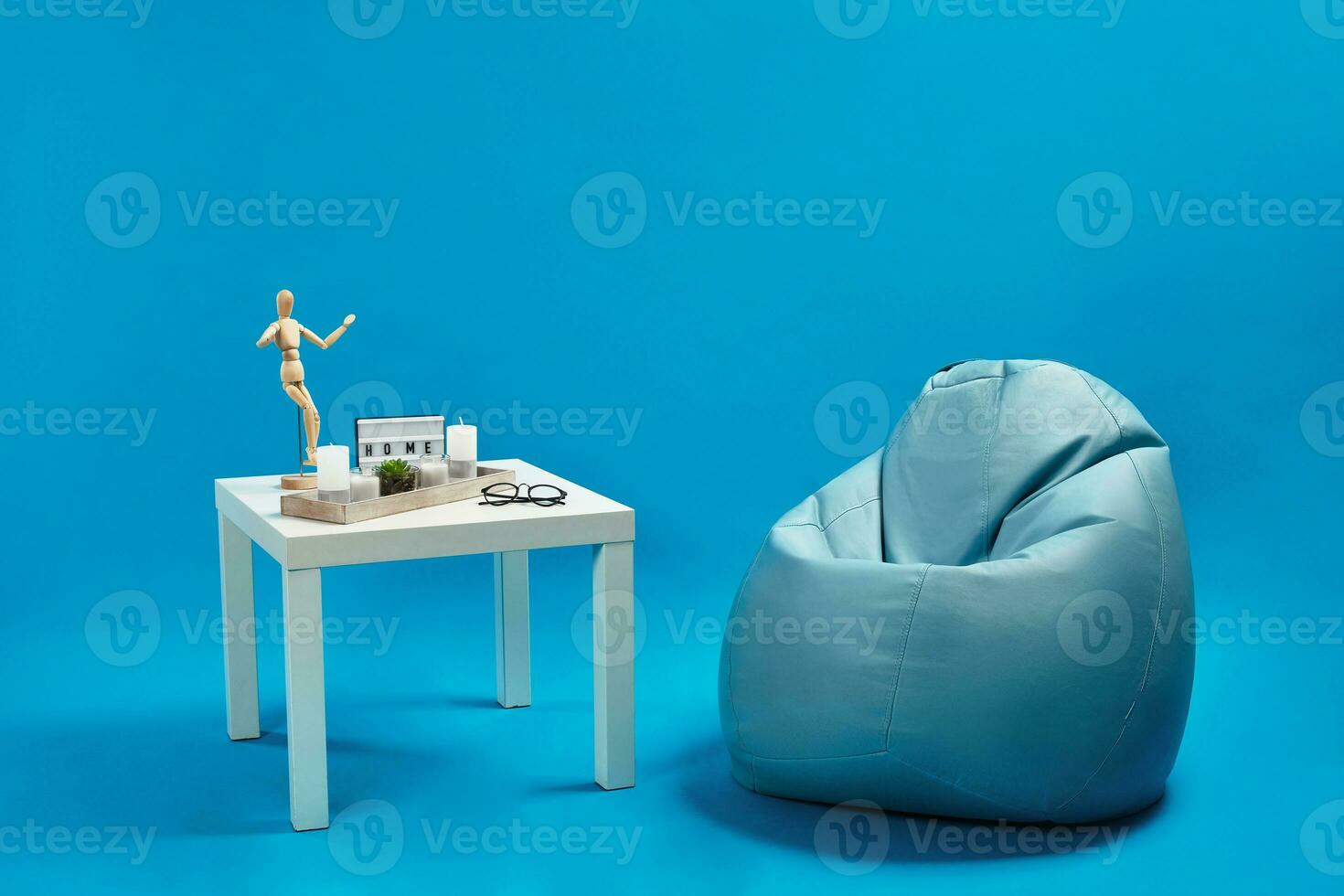 Turquoise bean bag, white coffee table with candles and succulent in glass pot, wooden figurine of human, glasses. Blue background. Copy space photo