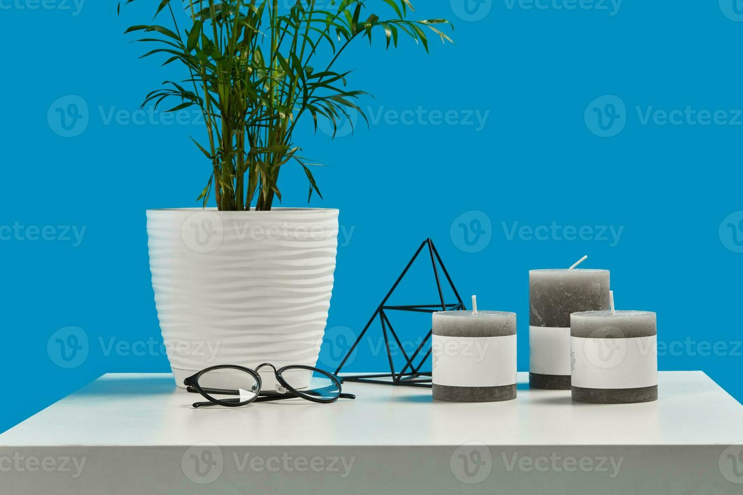 White coffee table with green flower in pot, three candles, decorative iron triangle and black rimmed glasses. Blue background. Close up, copy space photo