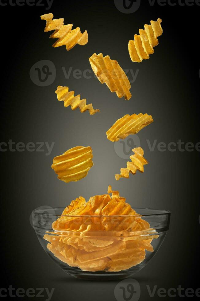 Grooved potato crisps falling down in glass bowl against a black background with copy space for text, images. Crispy chips. Advertising. Close-up. photo