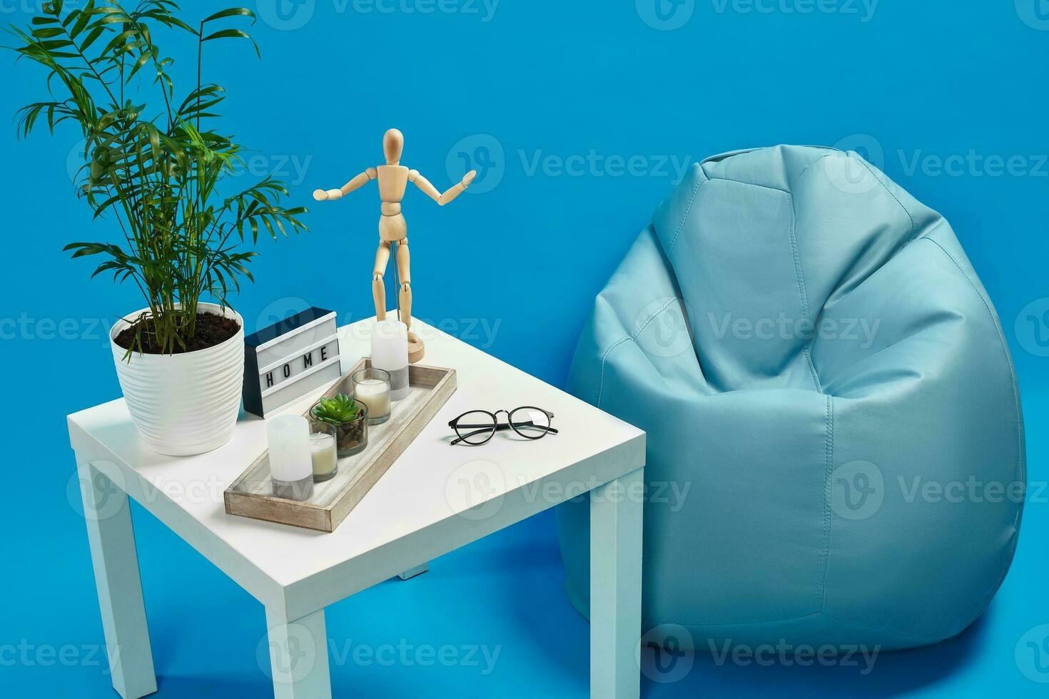 Bean bag, white table with green flower in pot, candles, succulent, wooden figurine of human, glasses, home decor element. Blue background. Close up photo