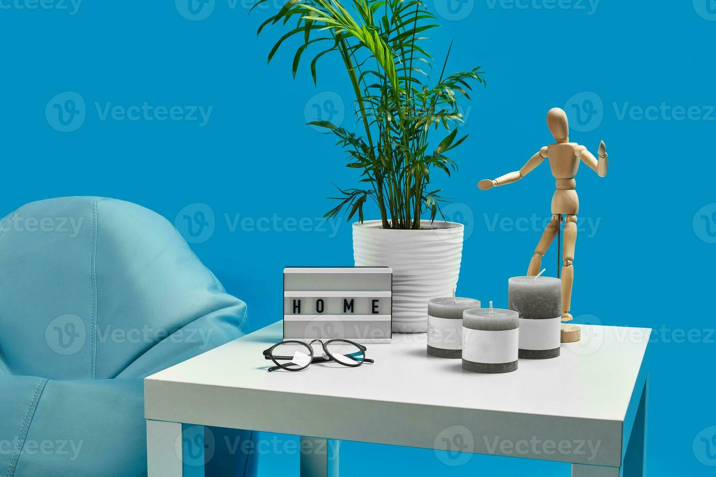 Turquoise bean bag, white coffee table with green flower in pot, three candles, wooden figurine of human, glasses. Blue background. Close up photo